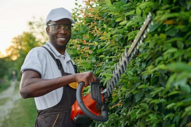 Jardinier africain souriant portant une combinaison spéciale avec des lunettes de protection travaillant avec du matériel de jardinage professionnel en plein air. Jeune homme utilisant un taille-haie au travail